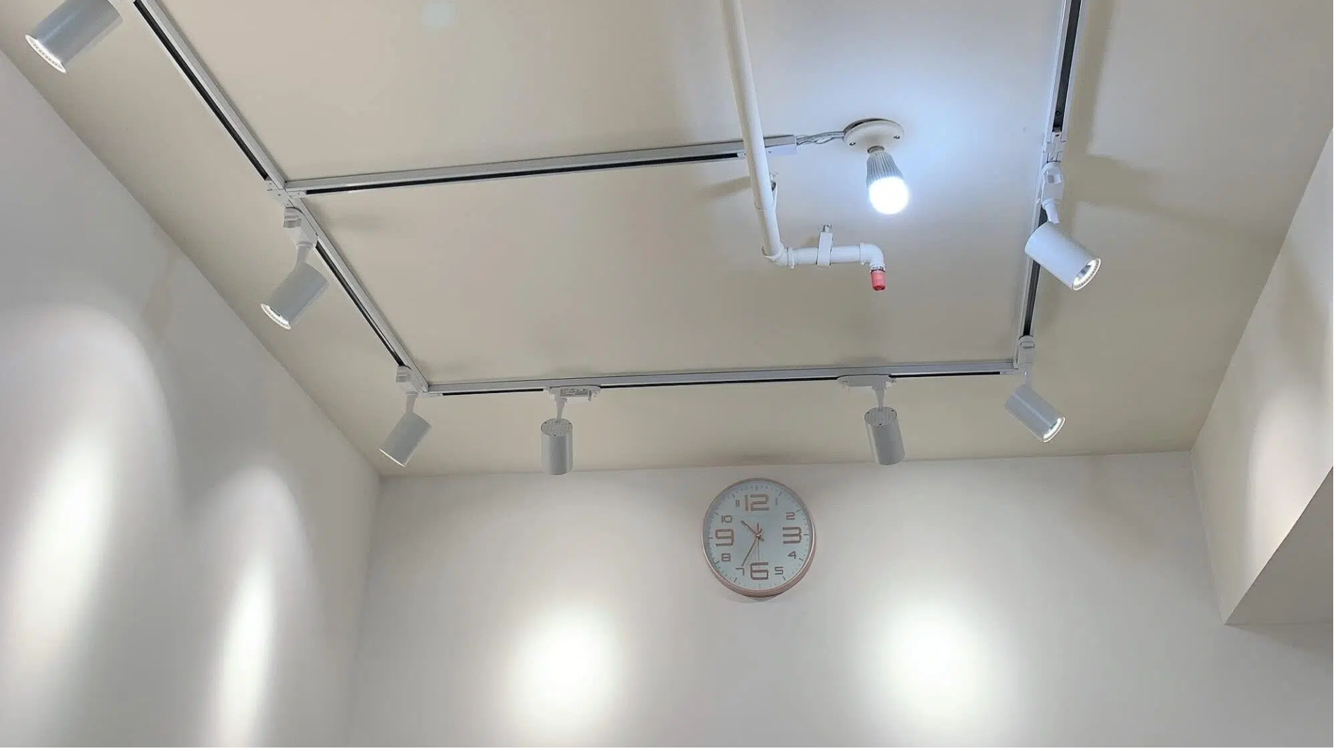 Square layout ceiling track lighting system installation with adjustable white spotlight heads and exposed conduit power feed for modern room illumination