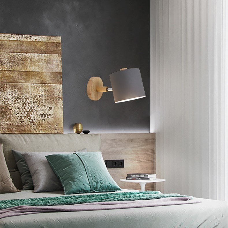Grey cylinder shade adjustable wall sconce with wood base, mounted above a bed, wireless, battery-powered.