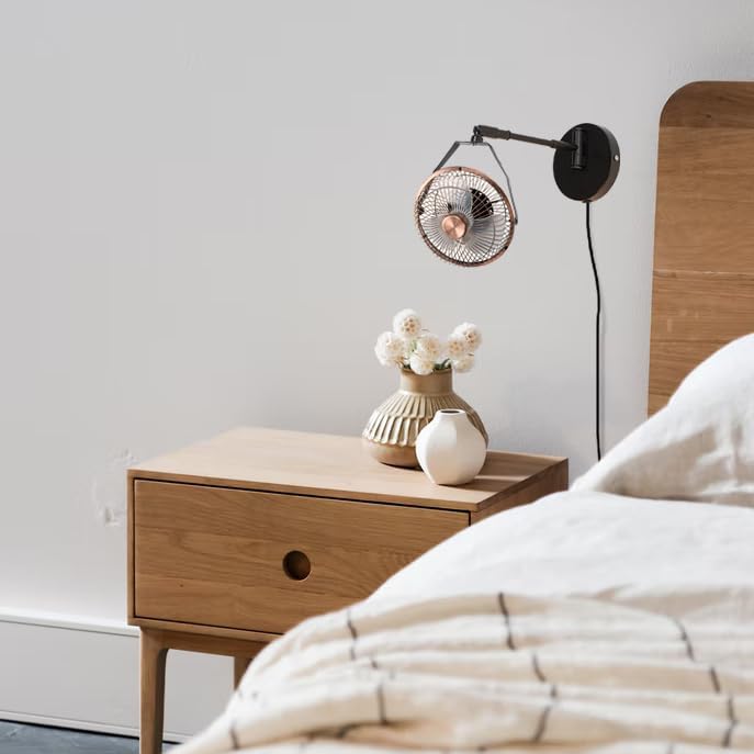 Industrial black wall mount fan with swing arm installed above a wooden bedroom nightstand